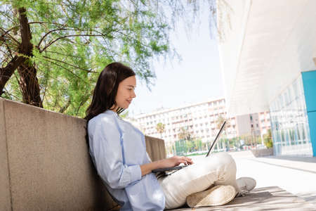 side view of smiling woman sitting with crossed legs on city street and typing on laptopの写真素材