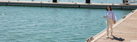 full length of brunette woman with paper cup walking along pier, banner