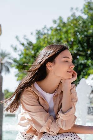 dreamy and positive brunette woman holding hand near face while sitting outdoors