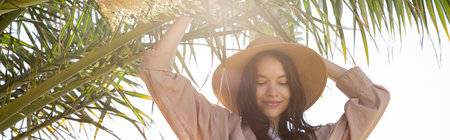 pleased brunette woman in straw hat standing near green palm tree, bannerの写真素材