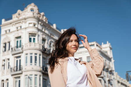 brunette woman standing on windy street near white buildings on blurred background