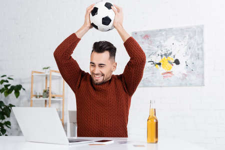 joyful bookmaker holding soccer ball above head near laptop and beerの写真素材