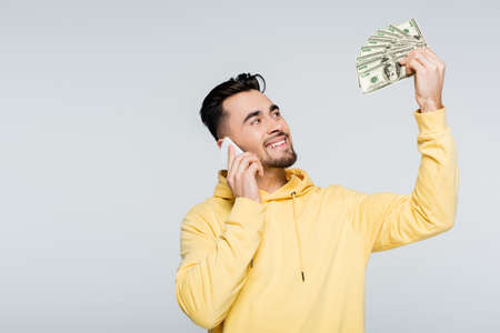 pleased bookmaker holding dollar banknotes while talking on cellphone isolated on grey