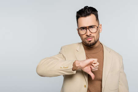 displeased critic in eyeglasses showing thumb down while looking at camera isolated on grey