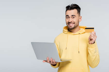 pleased man holding credit card and laptop while looking at camera isolated on grey