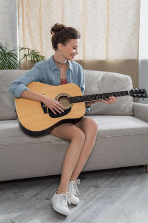 full length view of cheerful woman sitting on couch and playing acoustic guitar