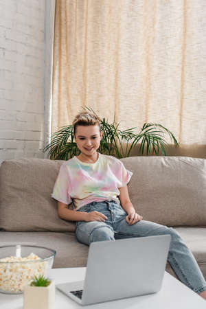 young and happy pansexual person with short hair sitting on couch and watching film on laptop