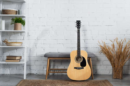 acoustic guitar, ottoman, vase with dry spikelets and rack with books and wicker baskets near white brick wallの写真素材