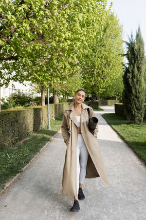 full length of woman in sportswear and trench coat standing with fitness mat in parkの写真素材