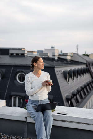 woman with clay cup sitting on rooftop near laptop and mobile phoneの写真素材