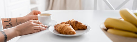 partial view of tattooed woman with coffee cup near croissants and blurred bananas, banner