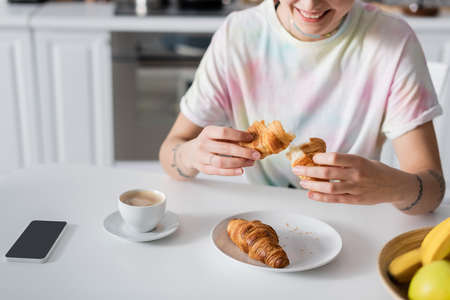 cropped view of smiling woman holding croissant near coffee cup and smartphone with blank screenの写真素材