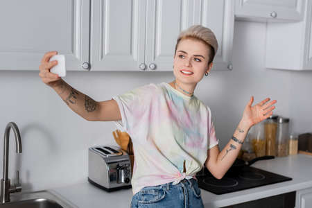 smiling tattooed woman taking selfie on cellphone in kitchen