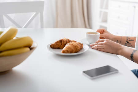 cropped view of tattooed woman near coffee cup, croissants, blurred bananas and smartphoneの写真素材
