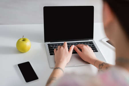 cropped view of blurred woman typing on laptop near smartphone with blank screen and ripe apple
