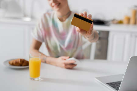cropped view of blurred woman with credit card near laptop, glass of orange juice and croissant