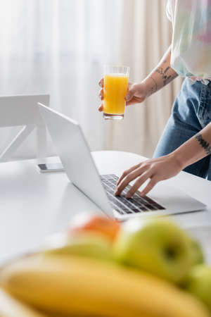 partial view of tattooed woman with orange juice typing on laptop near blurred fruits