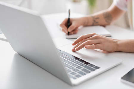 partial view of woman writing in blurred notebook near laptop