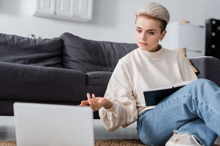 woman in white pullover sitting on floor with notebook and pointing at blurred laptop