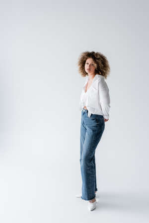 Curly woman in blouse and jeans looking at camera on white backgroundの写真素材
