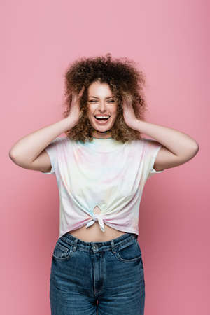Happy curly woman touching hair isolated on pink