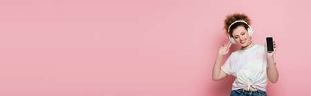 Cheerful woman in headphones showing smartphone on pink background, bannerの写真素材