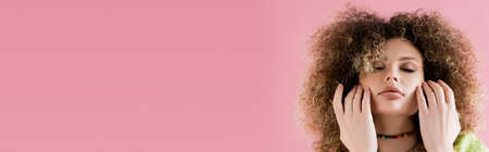 Young curly model with closed eyes touching face isolated on pink, bannerの写真素材
