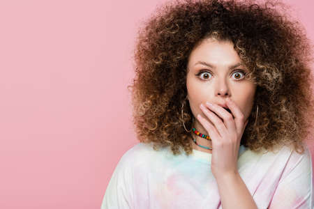 Portrait of shocked curly woman covering mouth isolated on pink