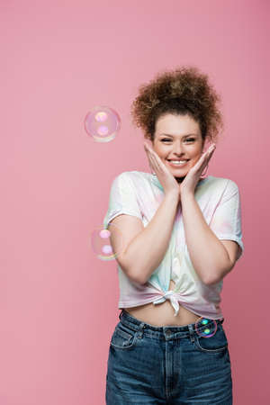 Positive woman in t-shirt posing near soap bubbles on pink backgroundの写真素材