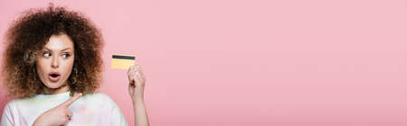 Curly woman pointing at credit card with finger isolated on pink, bannerの写真素材