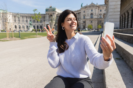 happy woman showing peace sign while taking selfie on smartphone in viennaの写真素材