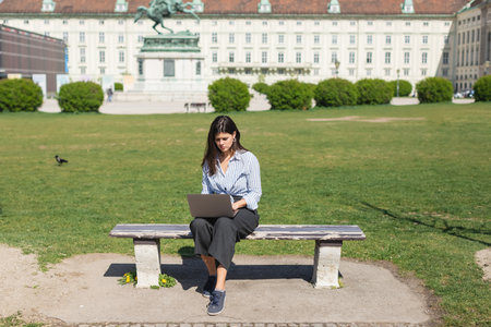 brunette woman sitting on bench and using laptop in green park of viennaの写真素材
