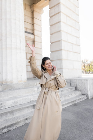 happy woman in beige trench coat waving hand while talking on smartphone on street near building of viennaの写真素材