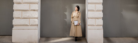 full length of happy woman in beige trench coat standing with laptop near arch, banner