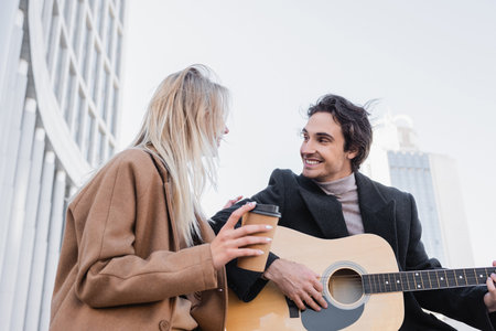 cheerful man looking at blonde woman with takeaway drink while playing acoustic guitar outdoorsの写真素材