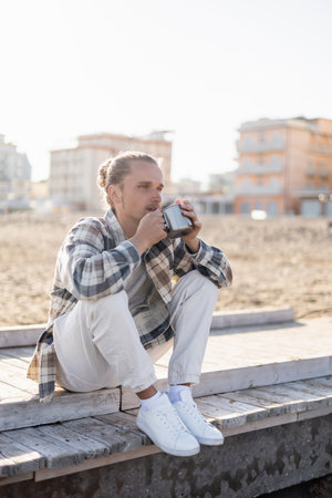 Long haired man holding cup while sitting on pier on beach in Italyの写真素材