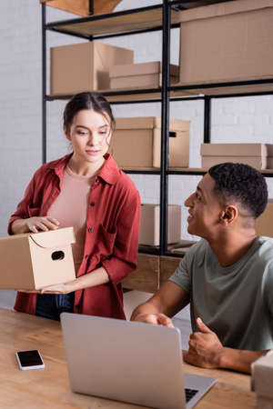 Smiling african american seller looking at colleague with package near devices in online web store
