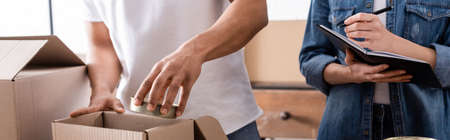 Cropped view of african american seller putting canned food in box near colleague with notebook in...