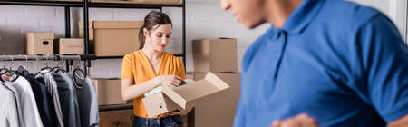 Proprietor holding box with clothes near blurred african american deliveryman in online web store, bannerの写真素材