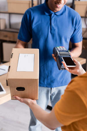 Cropped view of proprietor paying with smartphone and taking carton box from african american...