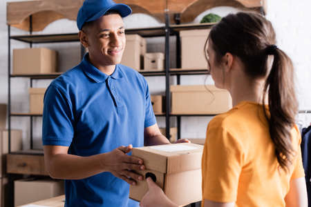 Smiling african american deliveryman taking carton box from proprietor in online web storeの写真素材