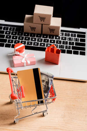 Credit card and money in toy shopping cart near laptop with small packages on tableの写真素材
