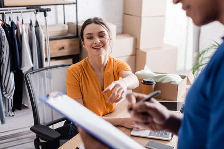 Smiling proprietor pointing at clipboard near blurred african american courier in online web storeの写真素材