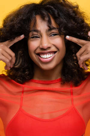 joyful african american woman pointing with fingers at her eyes isolated on yellow
