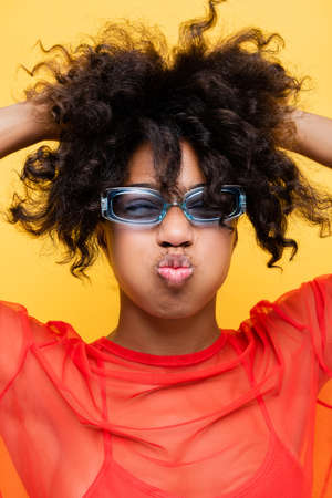 african american woman in trendy sunglasses pouting lips and touching wavy hair isolated on yellow