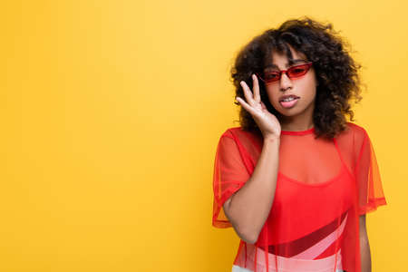 trendy african american woman in red top adjusting sunglasses isolated on yellow