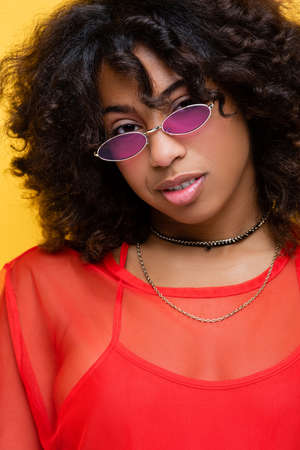 brunette african american woman in necklaces and trendy sunglasses smiling at camera isolated on yellowの写真素材