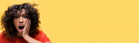 thrilled african american woman with open mouth touching face isolated on yellow, bannerの写真素材