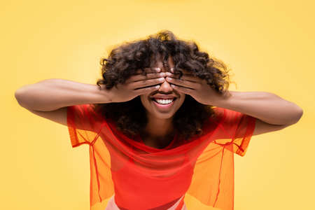happy african american woman in summer top covering eyes with hands isolated on yellowの写真素材
