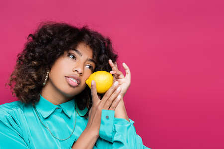pretty african american woman looking away while posing with whole lemon isolated on pinkの写真素材
